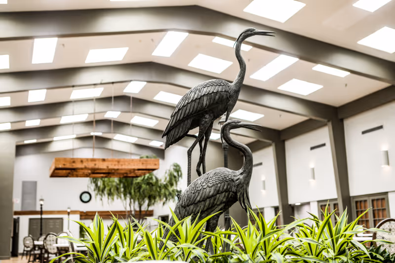 Indoor common area with a high ceiling and skylights, featuring a decorative sculpture of two large birds standing among green plants. In the background, there are tables and chairs, a large wooden light fixture, and some indoor trees.