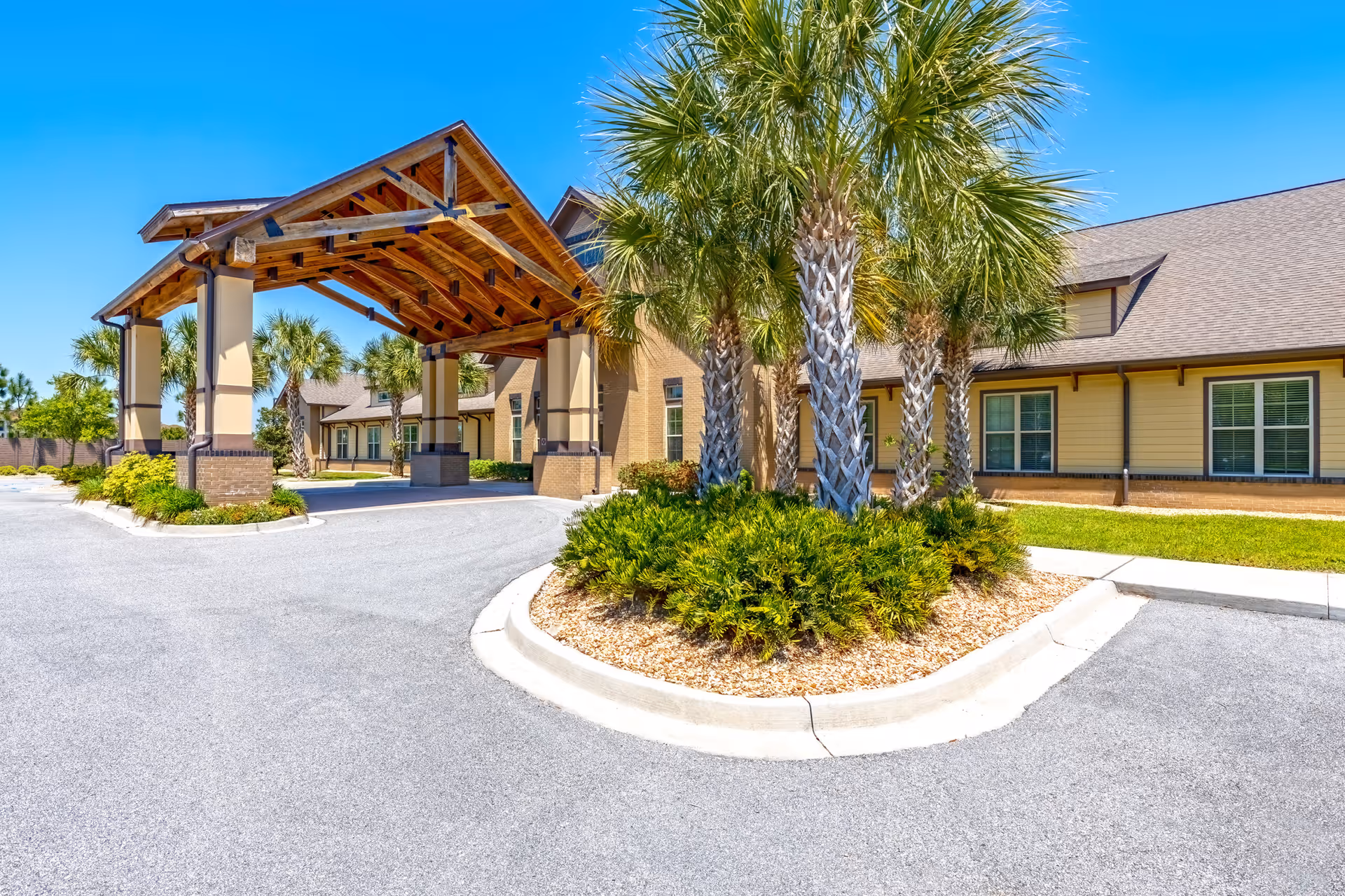 Entrance of a senior living facility with a large covered driveway supported by columns, surrounded by palm trees and landscaping under a clear blue sky.