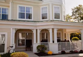 Exterior view of a light yellow two-story building with white trim, featuring a covered porch with white railings and decorative columns. There are shrubs and flowers planted along the front of the porch, and trees are visible in the background.