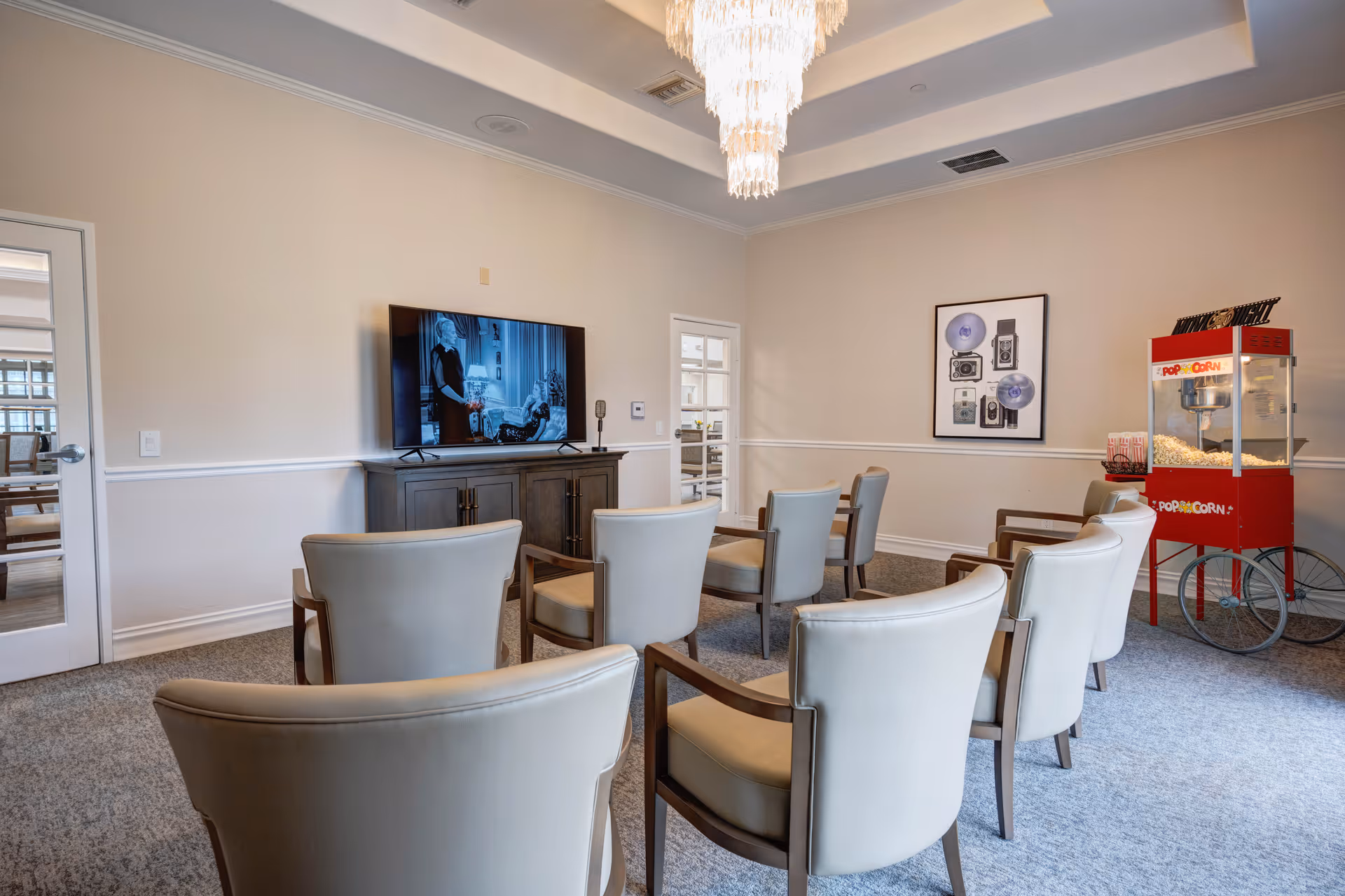 A small theater-style room with eight beige cushioned chairs arranged in rows facing a flat-screen TV mounted on a dark wooden cabinet. A red popcorn machine filled with popcorn is positioned in the corner of the room. The walls are light beige with white trim, and a framed picture of vintage cameras hangs on one wall. A chandelier with cascading crystals hangs from the ceiling.