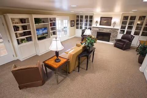 A spacious living room with beige carpet and cream-colored walls. The room features a yellow sofa, a brown armchair, a wooden side table with a lamp, and a black console table with a plant. There is a stone fireplace with a painting above it, flanked by built-in white bookshelves filled with books and decorative items. French doors with glass panes lead to another room, and recessed ceiling lights illuminate the space.