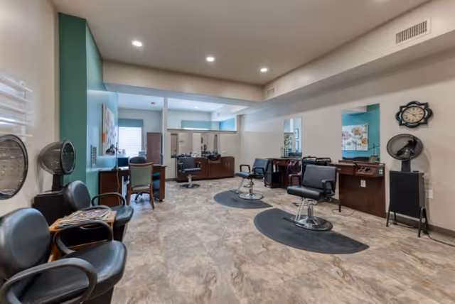 Interior view of a hair salon with multiple styling chairs, mirrors, hair dryers, and workstations. The room has a tiled floor, recessed ceiling lights, and a wall clock. There are chairs along the left side and a green accent wall with a window.