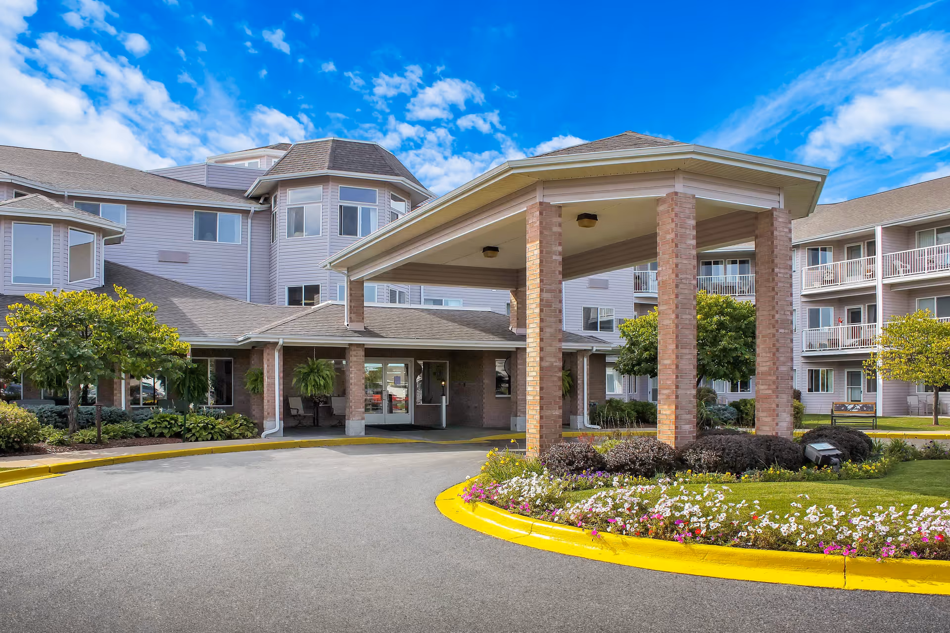 Front exterior view of Independence Village of Saginaw showing a covered entrance with brick pillars, a circular driveway, landscaped flower beds, and a multi-story building with balconies and windows under a blue sky with some clouds.