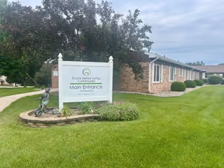 White sign reading "Evans Senior Living Community Main Entrance" on a grassy lawn in front of a single-story brick building.