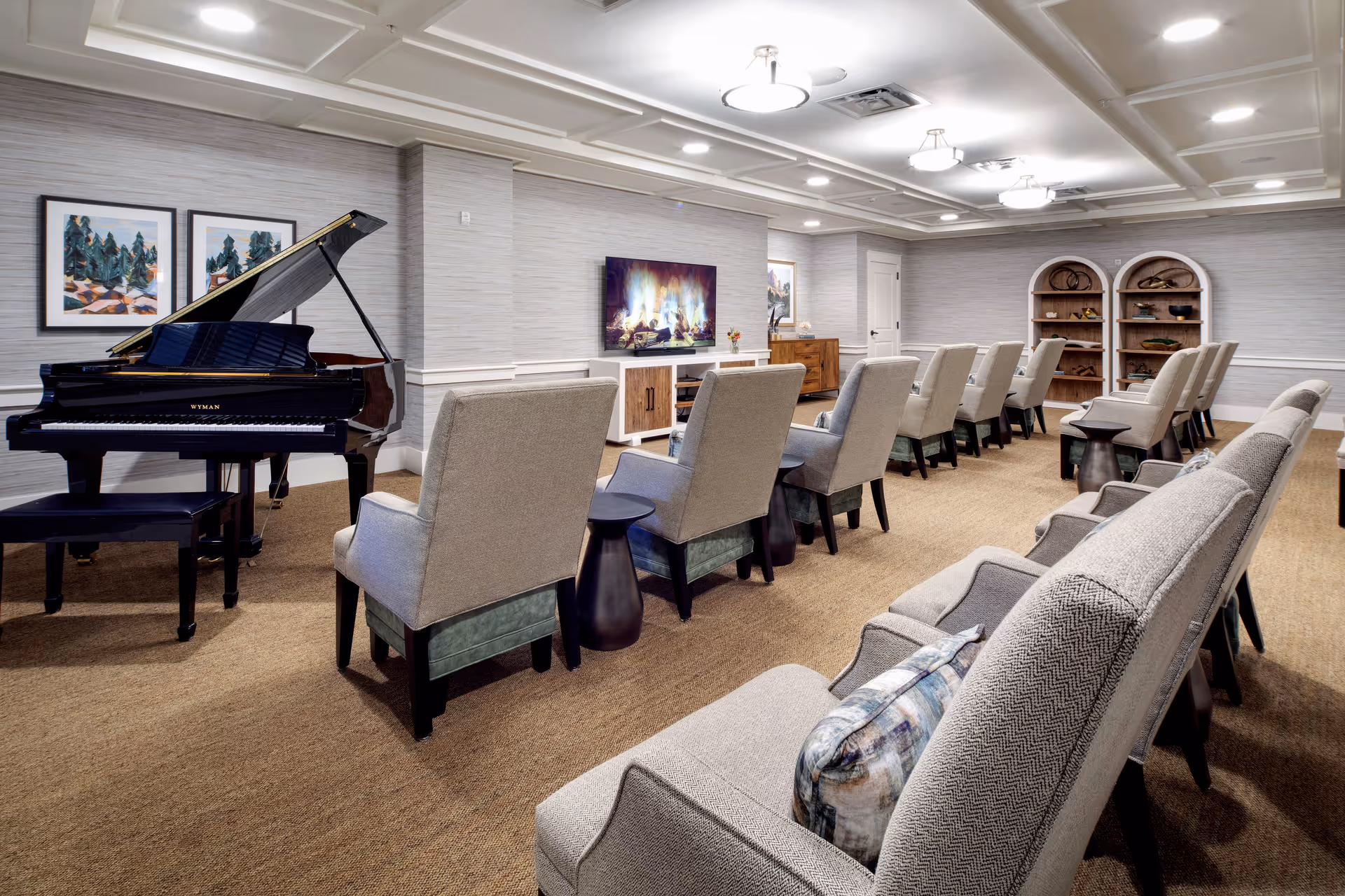A spacious, well-lit common room with rows of beige upholstered chairs facing a wall-mounted TV displaying a fireplace scene. To the left, there is a black grand piano with a matching bench. The walls are decorated with framed artwork and two arched shelves holding decorative items. The room has a carpeted floor and recessed ceiling lights.
