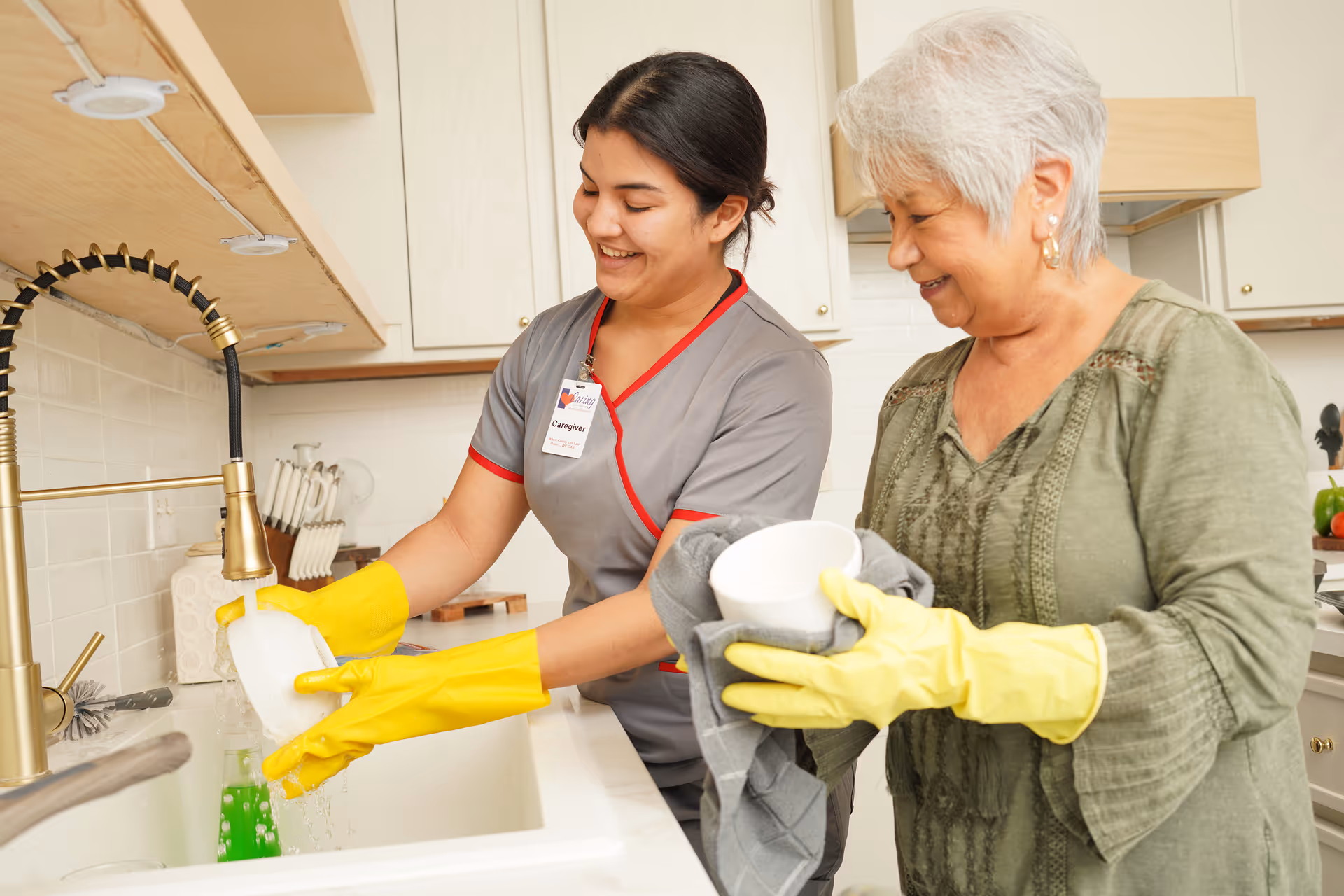 A caregiver and an elderly woman washing dishes together in a kitchen, both wearing yellow rubber gloves.