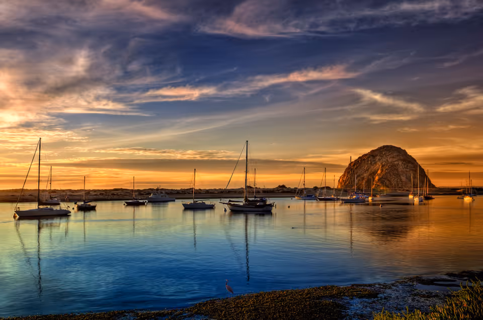 A serene coastal scene at sunset with several sailboats anchored in calm water and a large rocky formation in the background under a partly cloudy sky.