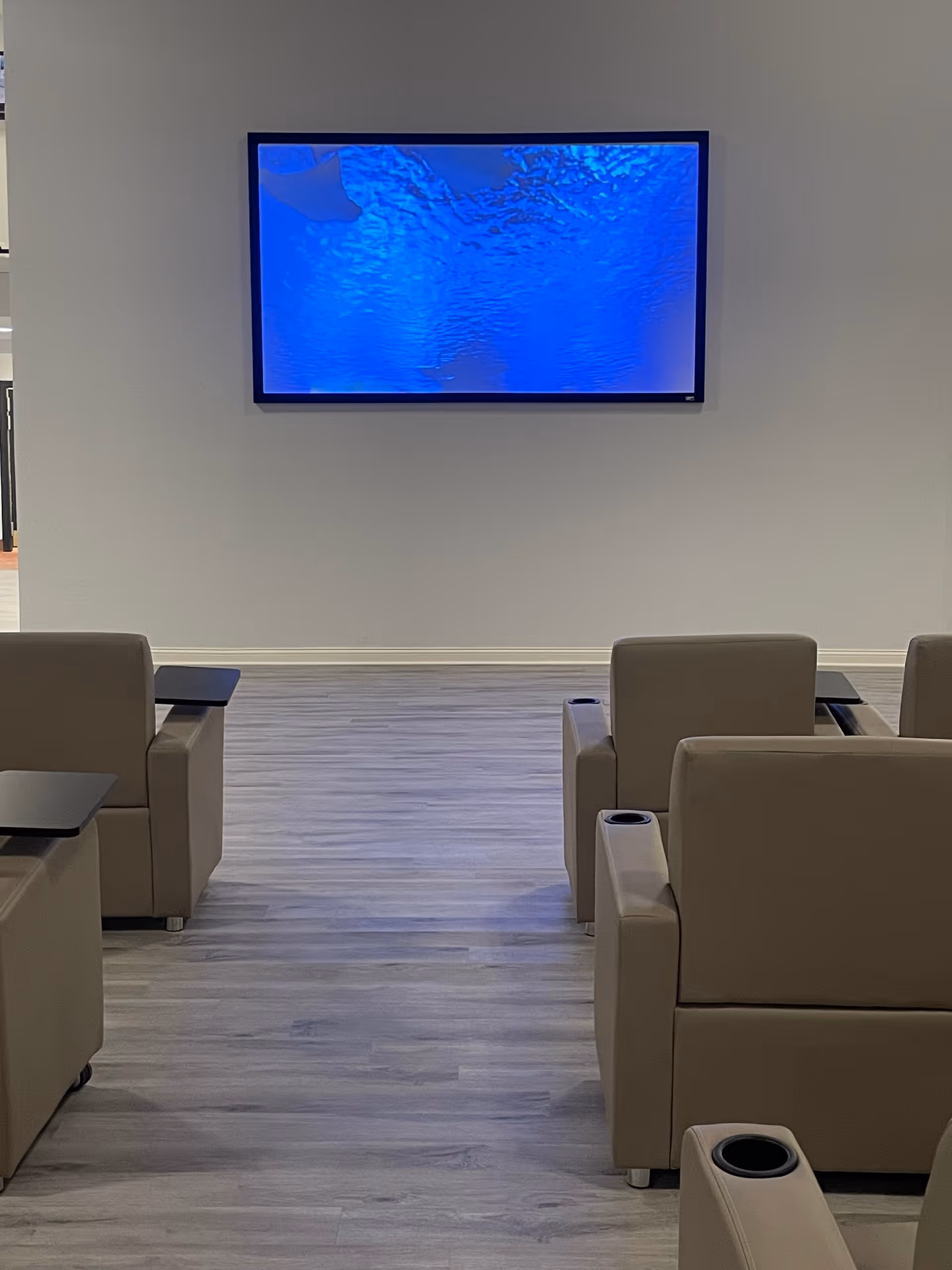 Beige recliner chairs with cup holders arranged facing a wall-mounted television displaying a blue screen in a common room.