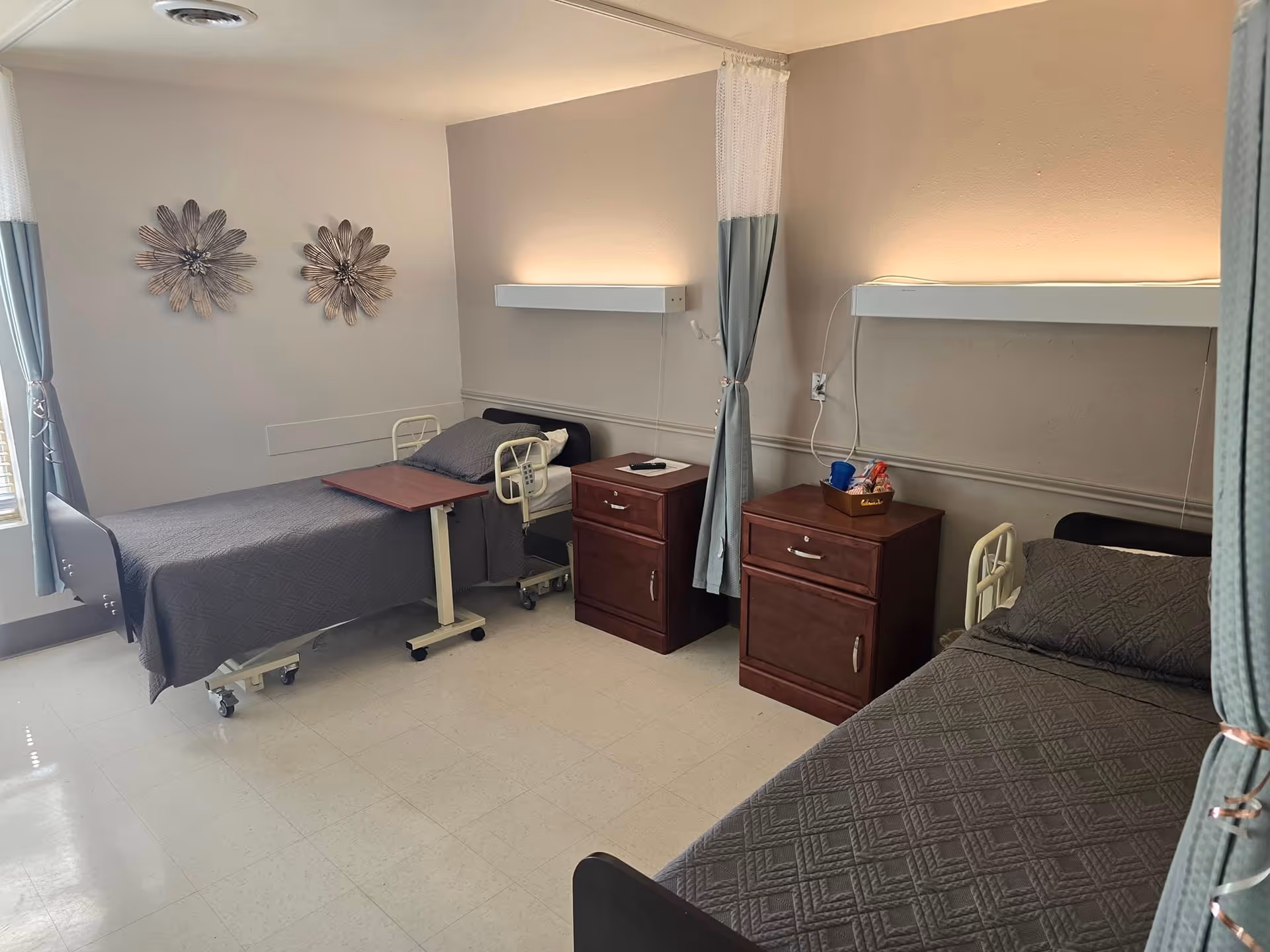 A shared patient room in a healthcare facility featuring two hospital beds with gray bedding, each accompanied by a wooden bedside cabinet. The room has light-colored walls with two decorative metal flower wall hangings, a window with curtains, and privacy curtains between the beds. Overhead lighting is mounted above each bed.