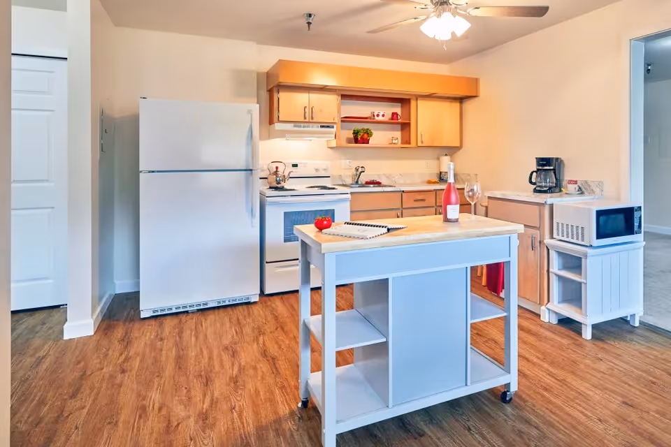 A bright kitchen area featuring a white refrigerator, white stove with oven, wooden cabinets, and a white kitchen island with shelves. On the island, there is a bottle of wine, a wine glass, a notebook, and a tomato. A microwave sits on a small white stand to the right, and a ceiling fan with lights is visible above.
