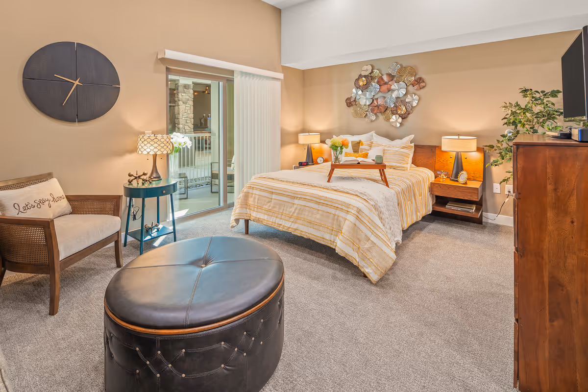A cozy bedroom with a bed covered in a striped yellow and white comforter, two bedside tables with lamps, a wooden dresser with a TV on top, a round black leather ottoman, a wooden chair with a cushion that says 'Let's stay home', a small round side table with a lamp, and a large wall clock. There is a sliding glass door leading to an outdoor patio area. The walls are painted beige and decorated with a metal floral wall art piece above the bed.