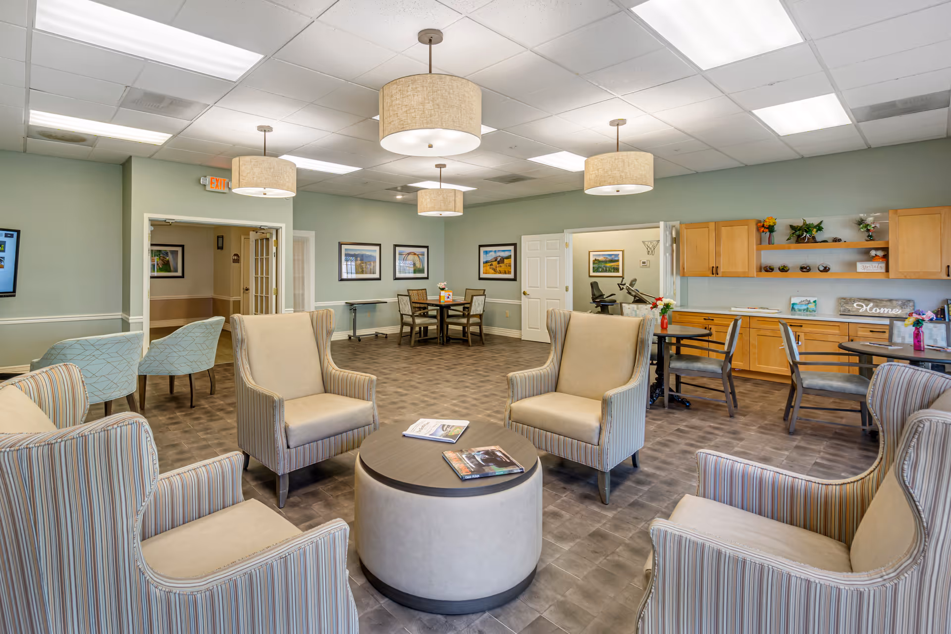 A spacious senior living common area with four beige and striped armchairs arranged around a round coffee table with magazines on it. The room has light green walls, several framed pictures, and multiple round tables with chairs. There are wooden cabinets and shelves with decorative items on the right side, and ceiling lights with beige lampshades hanging from a white tiled ceiling.