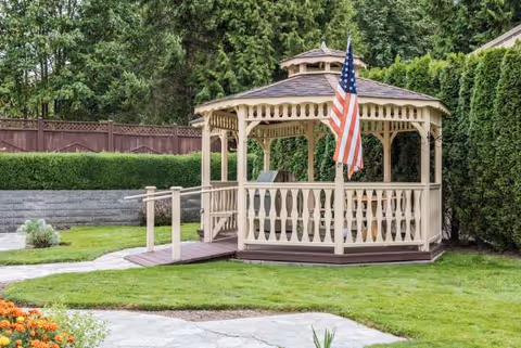 Cream gazebo with railing and American flag in a manicured backyard.