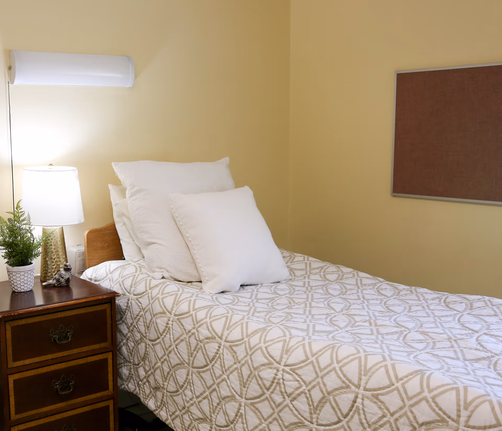 A neatly made single bed with white and beige patterned bedding and three white pillows against a beige wall. Next to the bed is a wooden nightstand with three drawers, a white table lamp, a small green potted plant, and a decorative bird figurine. A brown bulletin board is mounted on the wall above the bed.