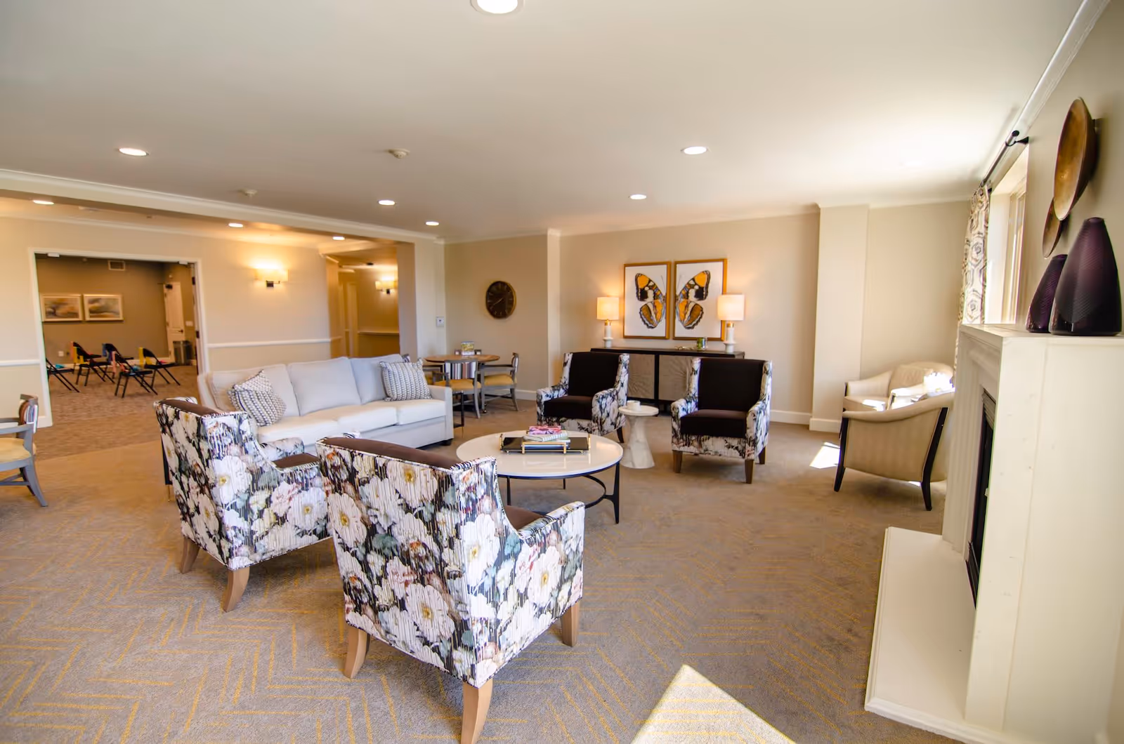 A bright and spacious living room area in a senior living facility with a white sofa, floral patterned armchairs, two dark armchairs, a round coffee table with books, and a fireplace. The room has beige walls, carpeted floor, and is decorated with butterfly artwork and table lamps. There is a window letting in natural light and a small dining table with chairs in the background.