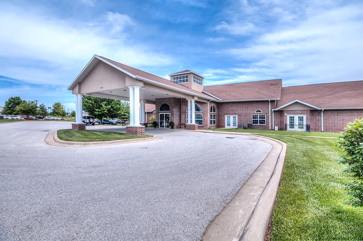 Front exterior view of a senior living facility named The Bungalows at Springfield East, featuring a covered entrance with white columns, brick walls, large windows, and a curved driveway with parked cars in the background under a partly cloudy sky.