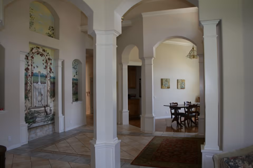 Interior view of a facility with white pillars and arches, tiled floor, a painted mural of a fountain on the left wall, and a dining area with a wooden table and chairs visible through an archway on the right.