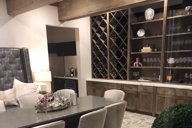 Interior view of a cozy lounge area featuring a gray cushioned chair, a table with a decorative tray holding flowers and glassware, and a wooden cabinet with wine storage and shelves displaying glassware and decorative items.