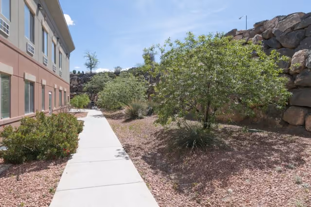 Sidewalk alongside a two-story building with desert landscaping, trees, and a rock retaining wall under a blue sky.