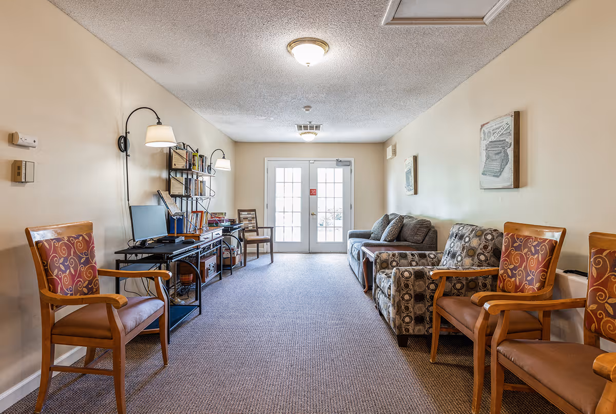 A long, narrow common area with beige walls and carpeted floor. The room has several wooden chairs with patterned cushions along the walls, a patterned armchair, and a gray sofa. There is a black metal shelving unit with books and a computer on a desk. Two wall-mounted lamps provide lighting, and double glass doors are at the far end of the room letting in natural light.