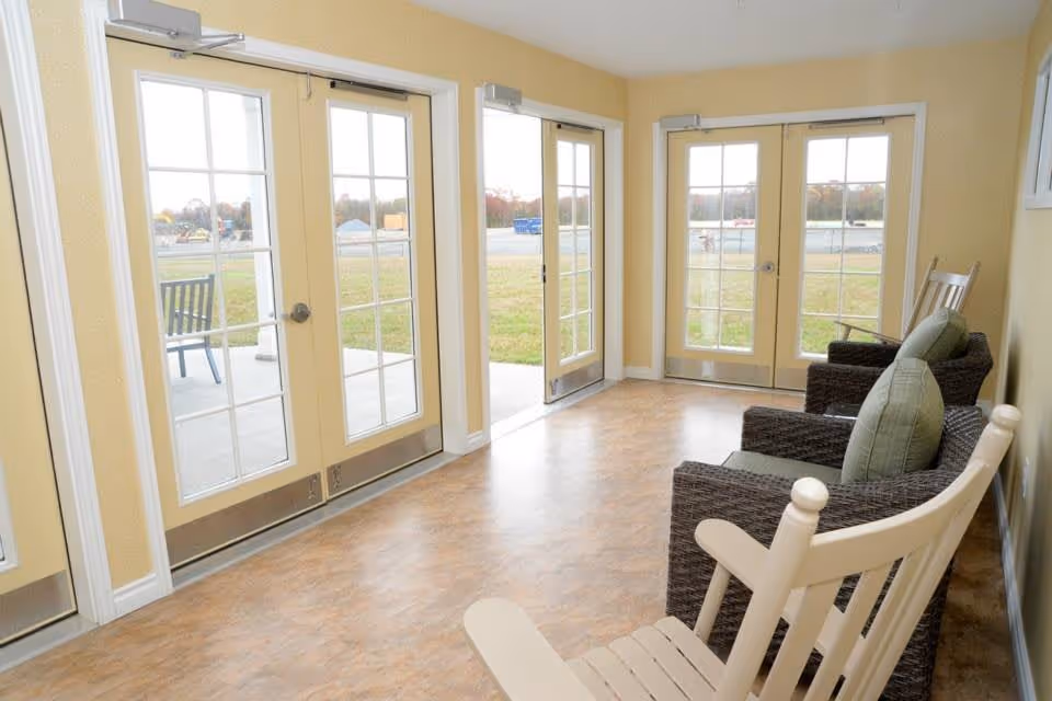 Bright sunroom with wicker and wooden rocking chairs arranged along a wall of glass French doors looking out to a lawn.