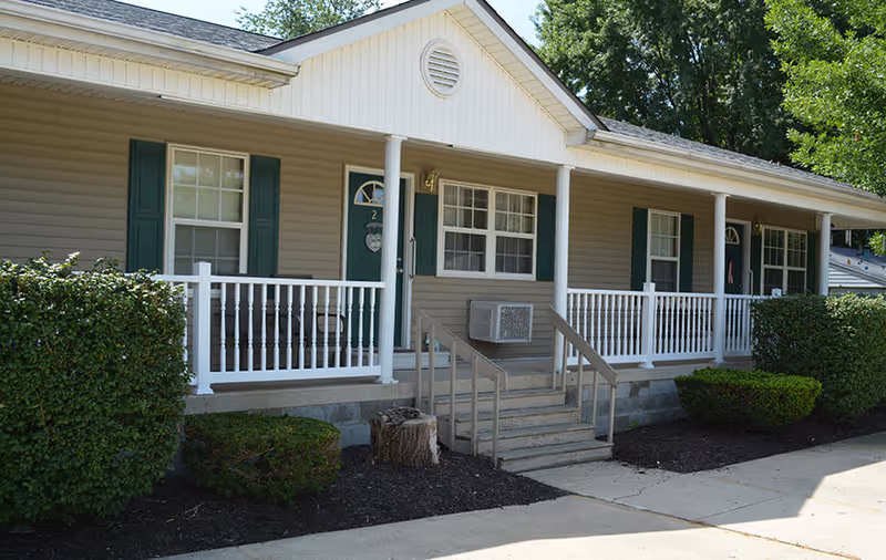 Single-story building front with a covered porch, white railing, steps leading to green doors and trimmed shrubs.