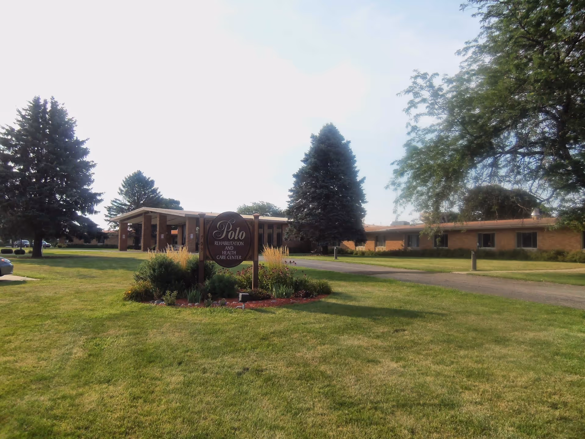 Exterior view of Polo Rehabilitation and Health Care Center building with a large green lawn, trees, and a sign displaying the facility name in front.