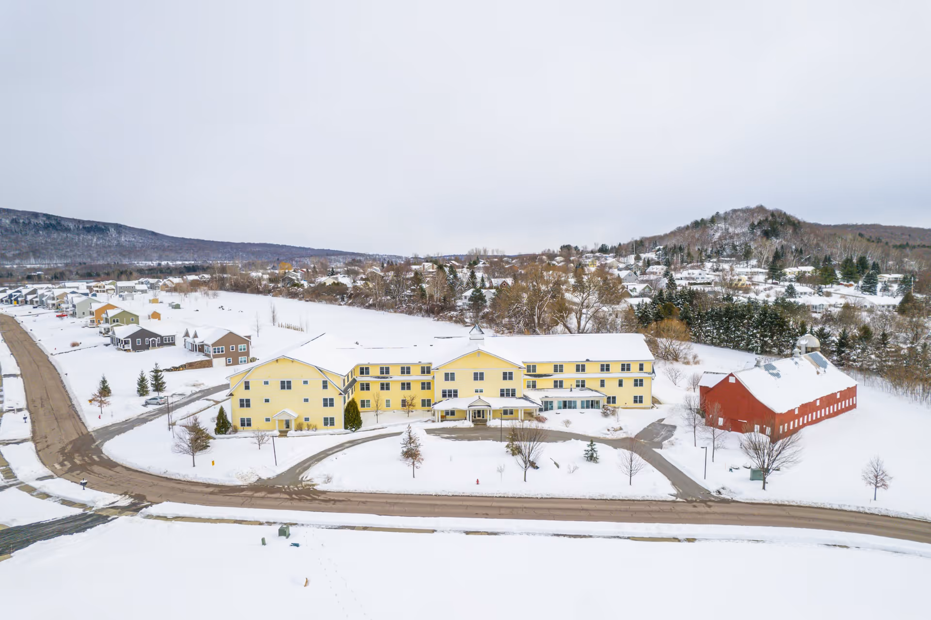 Aerial view of a large yellow senior living facility building surrounded by snow-covered grounds and roads, with a red barn nearby and a residential neighborhood in the background under a cloudy sky.