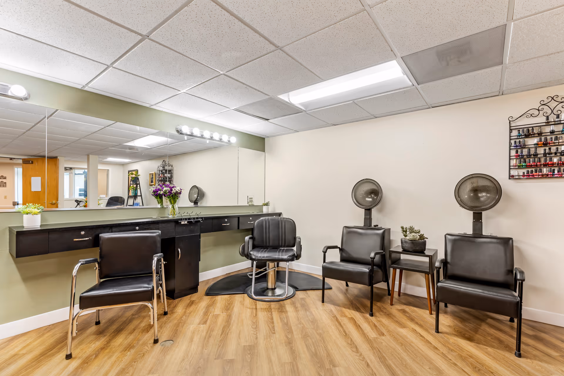 Interior view of a salon area in a senior living facility with black salon chairs, two hair dryers, a long mirror with lights above it, a small table with a plant, and a rack of nail polish on the wall.