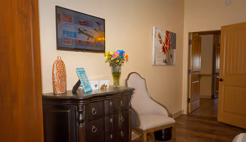 A wooden dresser topped with decorative vases, a framed sign and a vase of flowers beneath a wall-mounted TV, next to an upholstered chair and an open doorway.