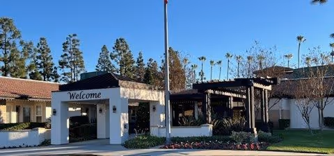 Front entrance of a senior living community with a 'Welcome' archway, flagpole, pergola, and landscaped grounds.