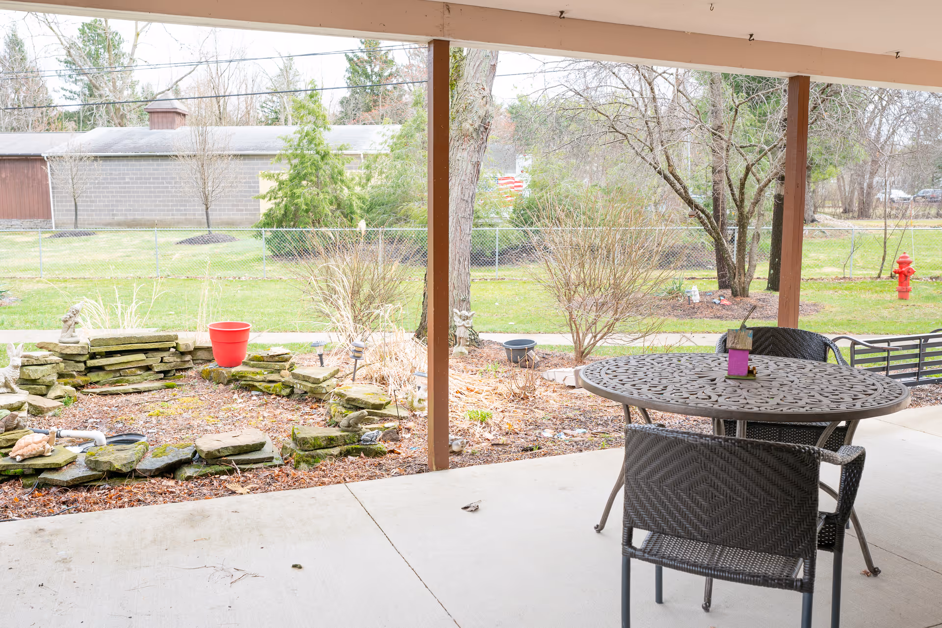 Covered patio with a round metal table and chairs overlooking a small rock garden, lawn, trees, and a chain-link fence.