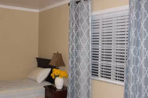 A tidy bedroom with a bed, nightstand, lamp, flowers, and patterned curtains.