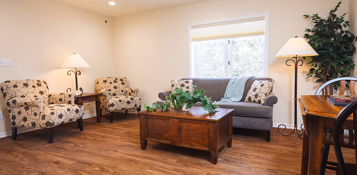 A cozy living room with two floral patterned armchairs, a gray sofa with floral pillows and a blue throw blanket, a wooden coffee table with a green leafy plant, two floor lamps, a wooden dining table with chairs, and a large window letting in natural light.