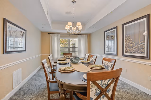 A formal dining room with a long wooden table set with place settings and upholstered chairs beneath a chandelier, with a window and framed artwork.