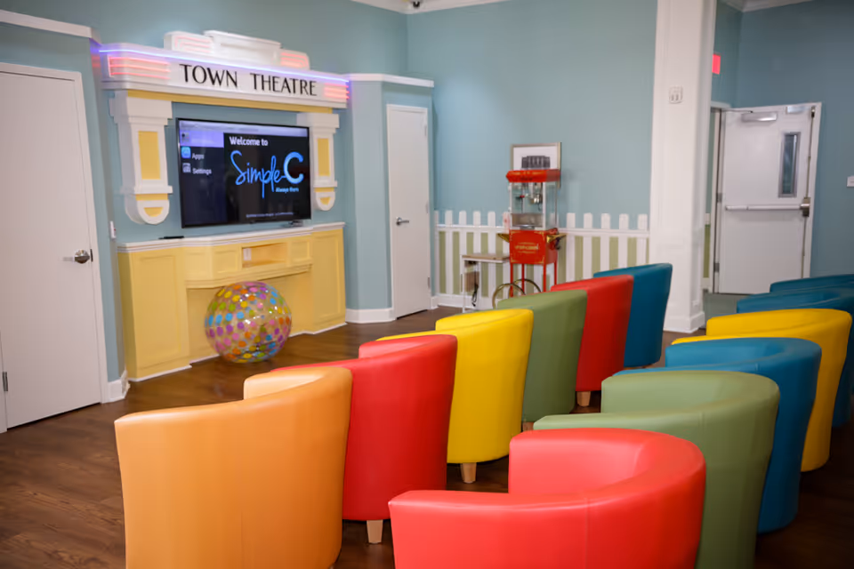 Colorful curved chairs arranged facing a wall-mounted TV beneath a 'Town Theatre' marquee in a pastel-colored activity room with a popcorn machine.