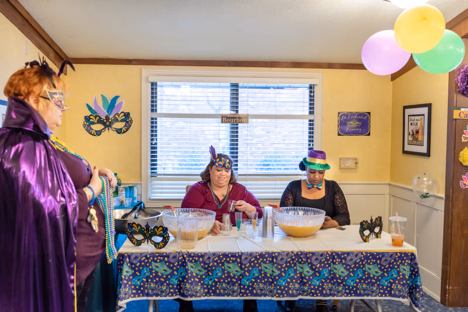Three women dressed in festive Mardi Gras attire are at a table indoors. Two women sit behind the table with large punch bowls and plastic cups, while a third woman stands to the left wearing a purple cape and mask. The table is covered with a colorful Mardi Gras-themed tablecloth and decorated with masks. Balloons are hanging from the ceiling, and a window with blinds is in the background.
