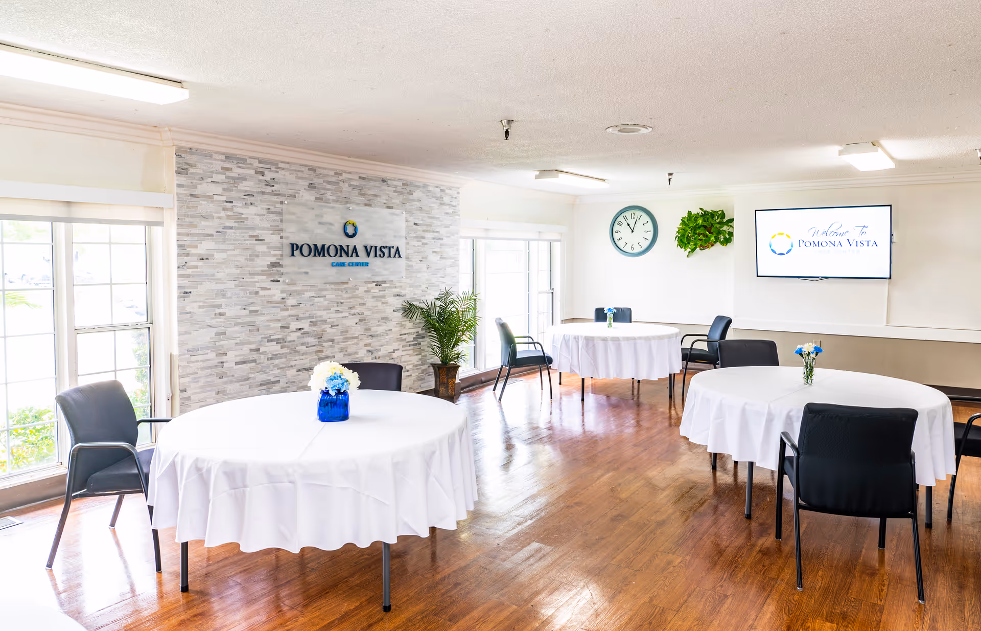 Bright dining room with round tables covered in white tablecloths, each adorned with small floral centerpieces. Black chairs surround the tables. Large windows let in natural light, and a wall features a sign that reads 'Pomona Vista Care Center.' A clock and a mounted TV screen displaying 'Welcome to Pomona Vista' are also visible on the walls.