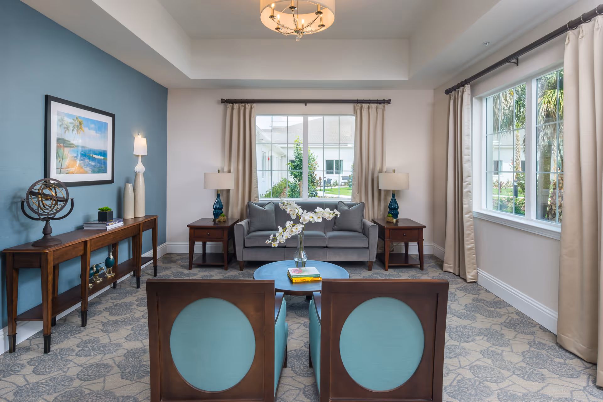 A bright and inviting living room with a gray sofa centered between two wooden side tables with blue glass lamps. In front of the sofa are two wooden chairs with light blue cushions and a round coffee table with a vase of white orchids and books. The room has large windows with beige curtains letting in natural light, a blue accent wall with a framed beach painting, and a wooden console table decorated with books and decorative items.