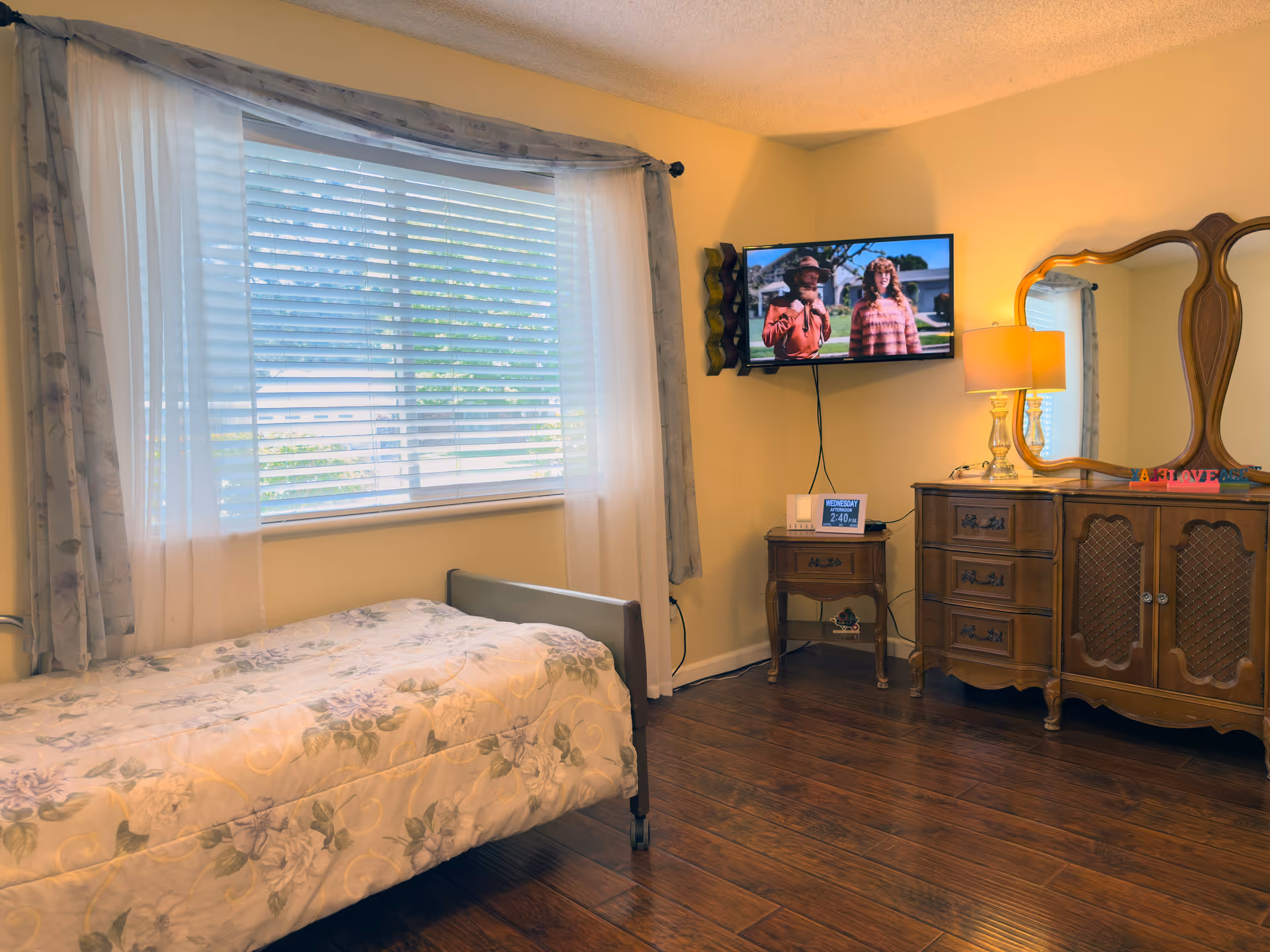 A cozy bedroom in a senior living facility with a single bed covered in a floral quilt near a large window with sheer and patterned curtains. On the opposite wall, a flat-screen TV is mounted above a wooden nightstand with a digital clock and a lamp. Next to the nightstand is a vintage wooden dresser with a large mirror and another lamp, creating a warm and inviting atmosphere.