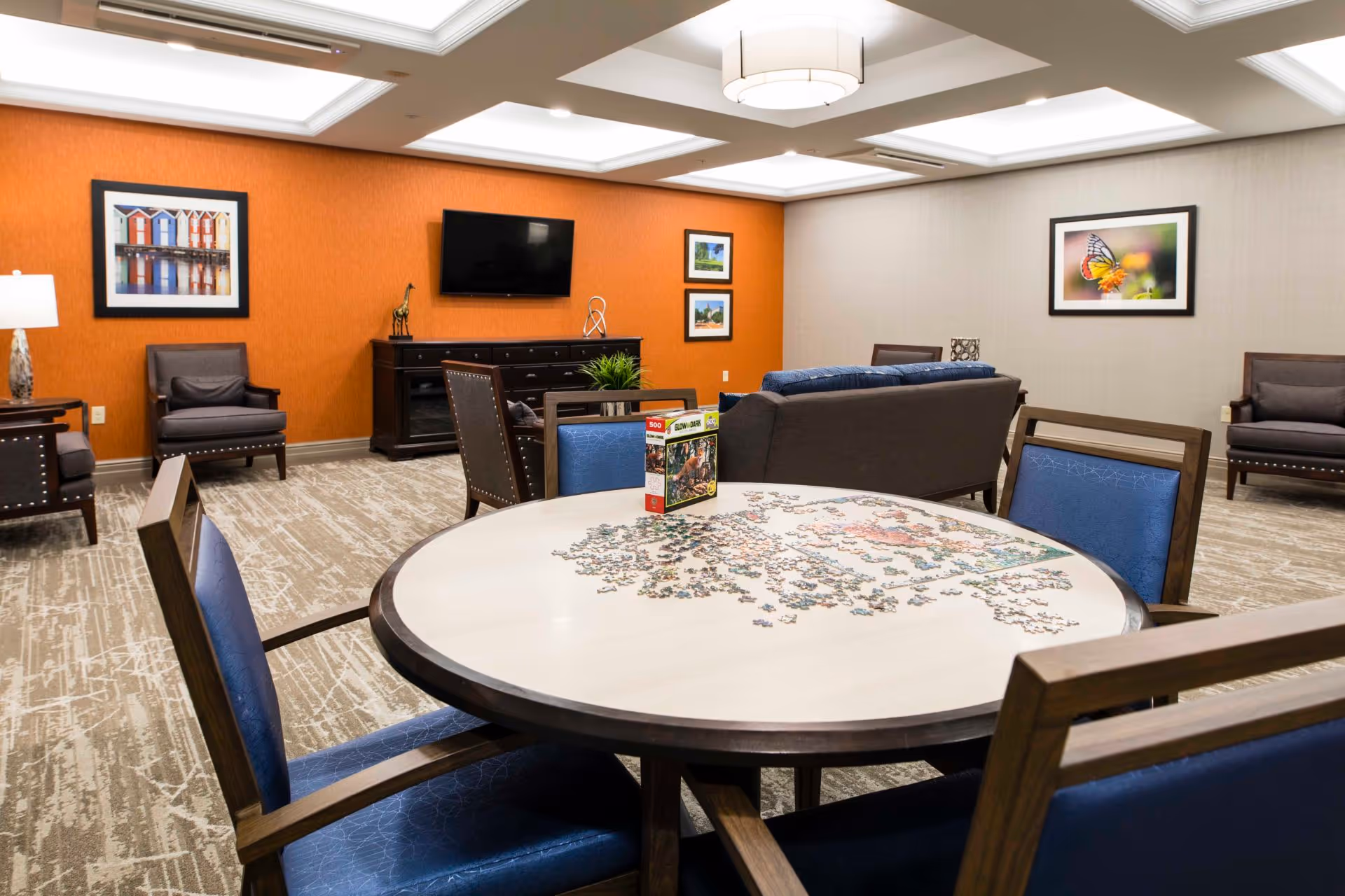 Communal activity room with a round table covered in a jigsaw puzzle, blue chairs, sofas, a TV on an orange accent wall, and framed artwork.
