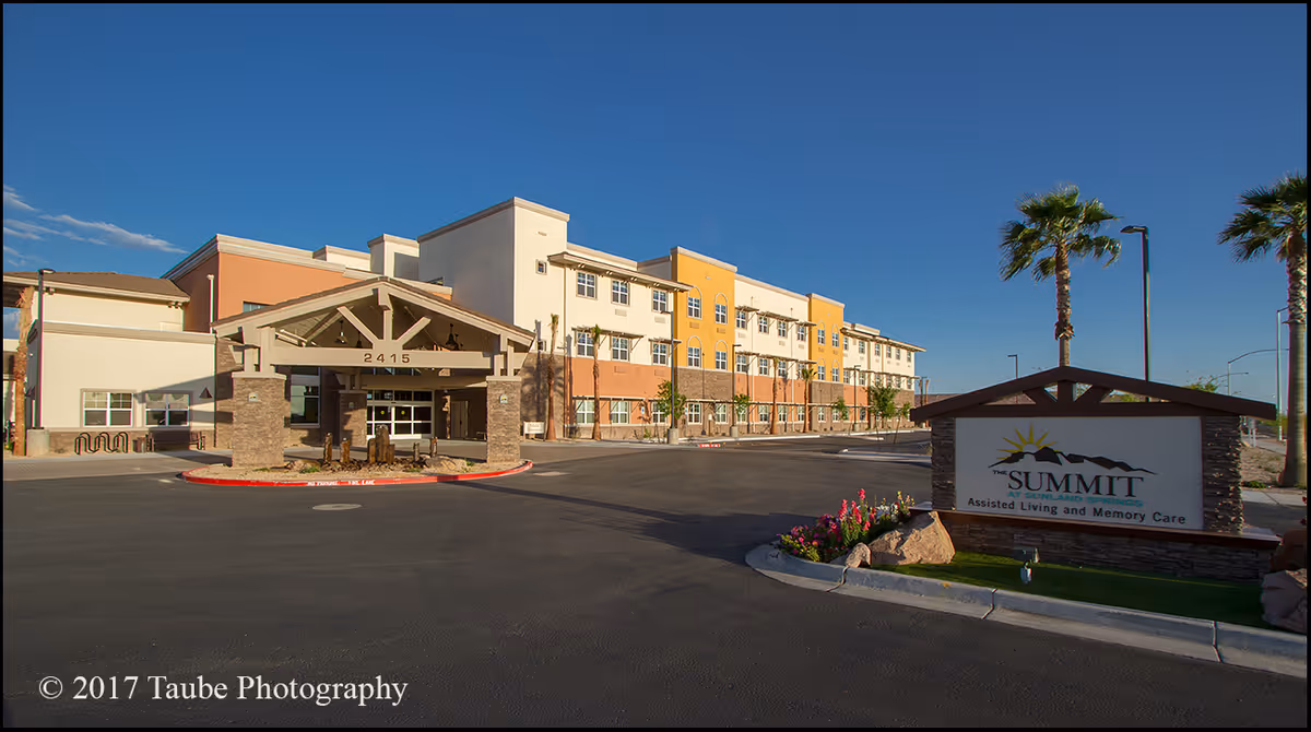 Exterior view of The Summit At Sunland Springs, a multi-story assisted living and memory care facility with a covered entrance, palm trees, and a sign displaying the facility's name and services.