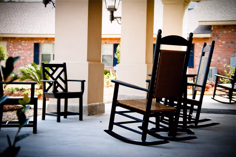 A covered outdoor patio area with several black wooden rocking chairs and armchairs arranged on a concrete floor. The background shows a brick building with windows and some landscaping including small plants and shrubs.