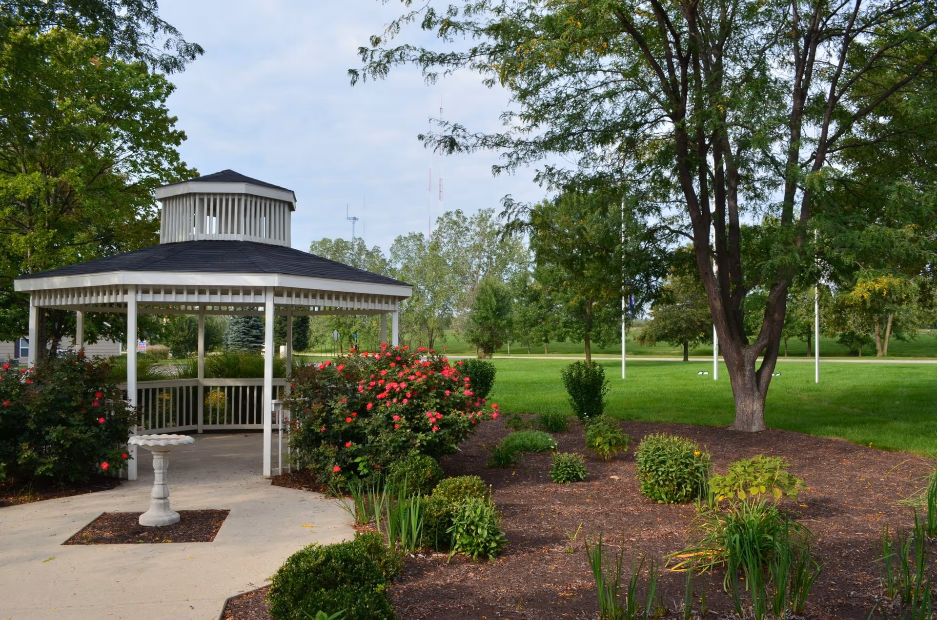 A white gazebo with a black roof situated in a landscaped garden area with blooming pink flowers, green shrubs, and a birdbath on a concrete path. Tall trees and a grassy lawn are visible in the background under a partly cloudy sky.