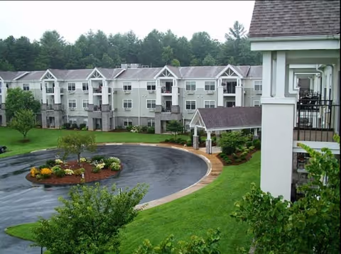 View of a multi-story residential building with balconies surrounding a circular driveway and landscaped garden area with trees and shrubs, set against a backdrop of dense forest.