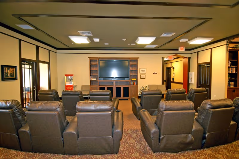 A cozy common room with multiple brown leather recliners arranged in rows facing a large flat-screen TV mounted on a wooden entertainment center. The room has beige walls with dark trim, a patterned carpet, and ceiling lights. There is a popcorn machine on the left side near the wall and an exit sign above a doorway on the right.