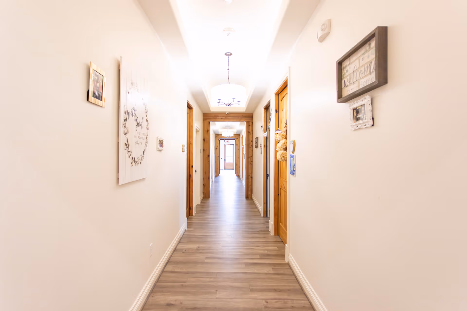 A brightly lit hallway with light-colored walls and wood flooring. Several wooden doors line both sides of the hallway, and decorative wall hangings including framed pictures and signs are visible. A ceiling light fixture is centered above the hallway, and a door with glass panels is visible at the far end.
