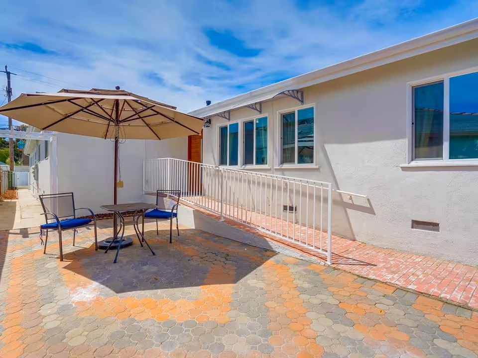 Outdoor patio area with a table, two chairs with blue cushions, and a large beige umbrella providing shade. The patio is paved with hexagonal tiles in shades of orange and gray. A white building with several windows and a white railing along a brick ramp is visible in the background under a blue sky with some clouds.