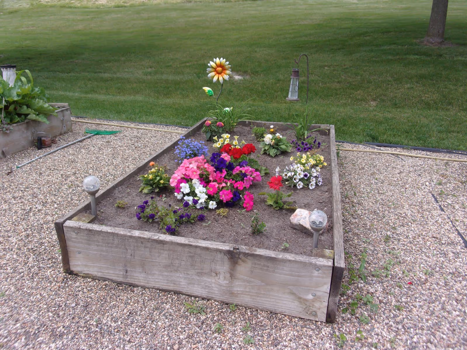 A raised garden bed filled with colorful flowers including pink, purple, red, white, and yellow blooms, surrounded by gravel and green grass in the background. There are garden decorations including a metal flower and a hanging lantern.