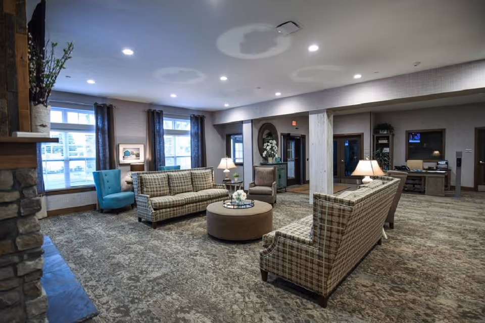 A spacious and well-lit living room area in a senior living facility with patterned carpet, two plaid sofas facing each other, a round ottoman in the center with a decorative tray, a blue armchair near the windows, and a stone fireplace on the left. The room has large windows with dark curtains, several table lamps, and a desk area in the background.